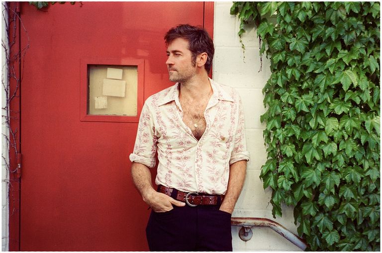 Singer-songwriter John Craigie leaning against a bright red door next to green ivy leaves, photo by Carly Montgomery.
